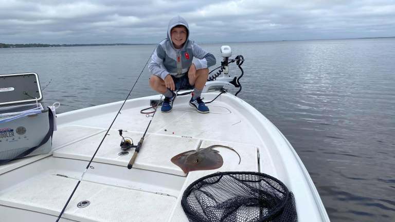 Person in hoodie squatting on boat deck with fishing gear and stingray.