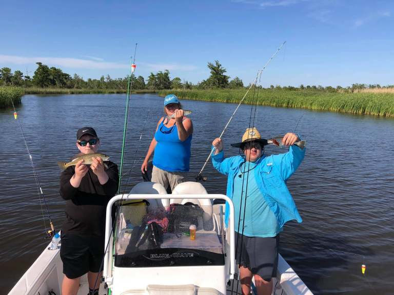 Three people on a boat holding fishing rods and caught fish in a lake.