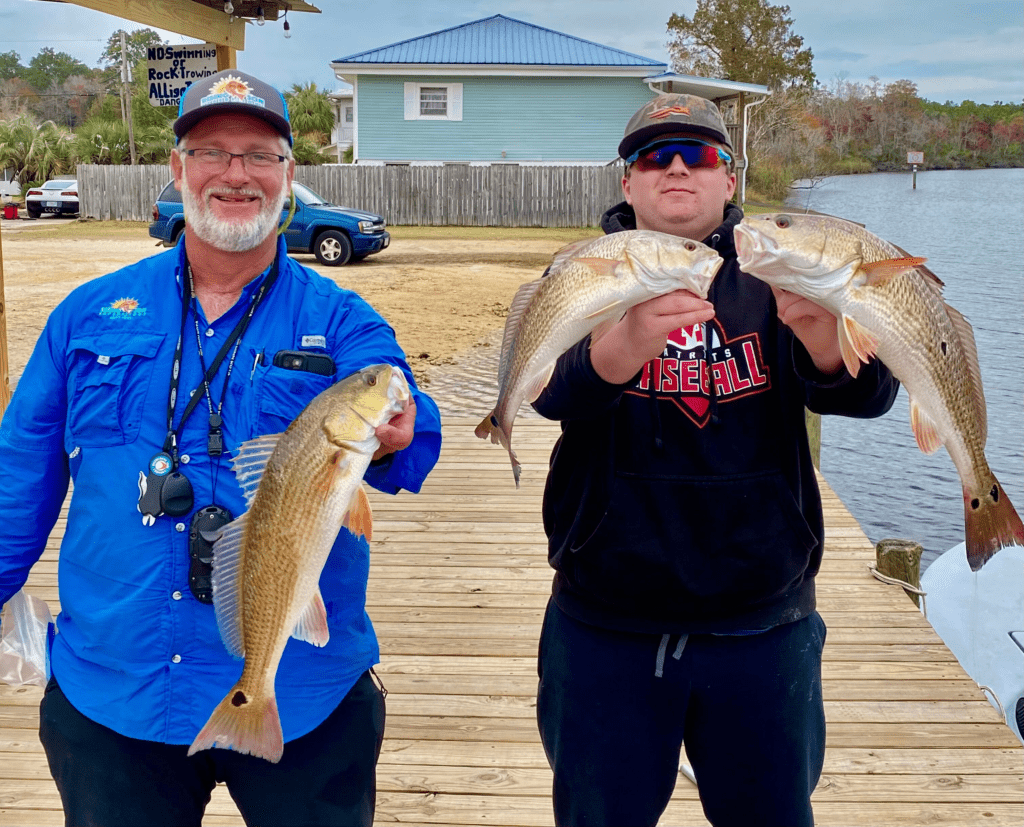 Two men smiling, holding large fish on a wooden dock near a river.