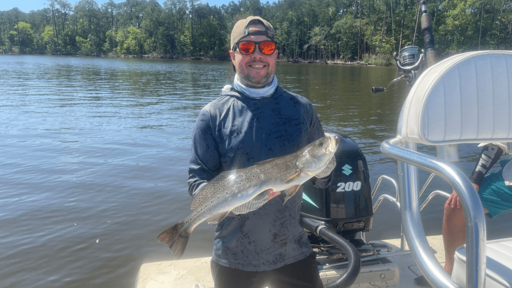 Person holding a fish on a boat with water and trees in the background.