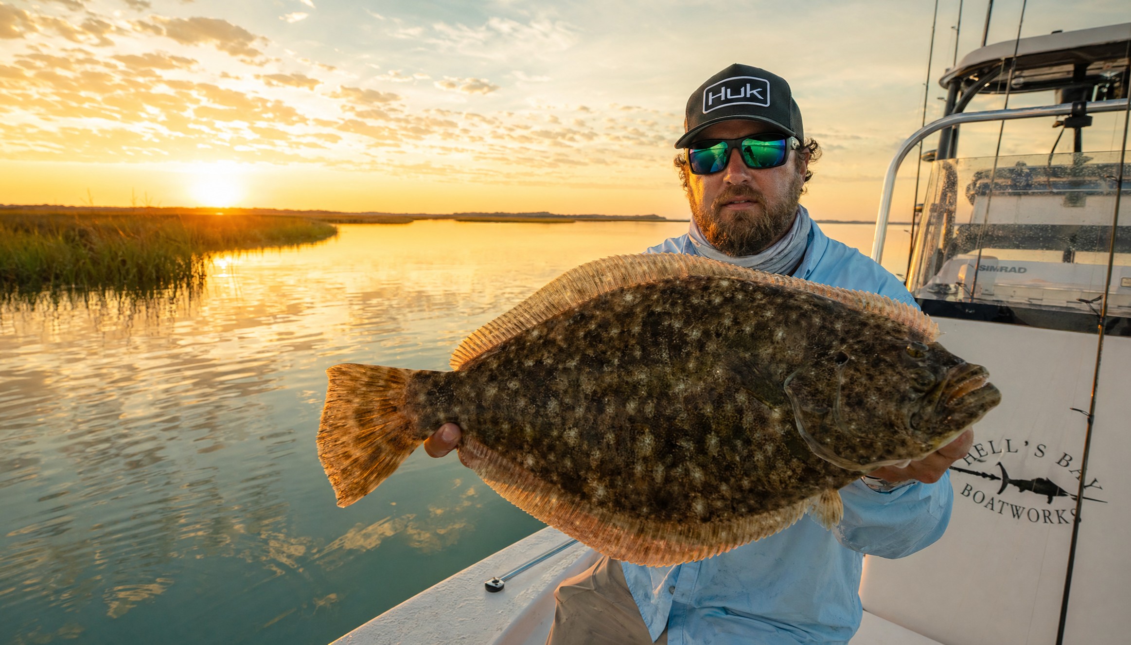 Man holding a large fish on a boat at sunset with calm water.