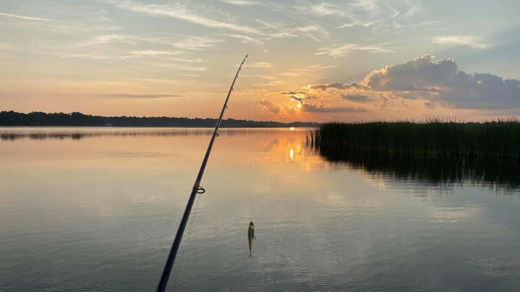 Fishing rod over calm lake at sunset with clouded sky.