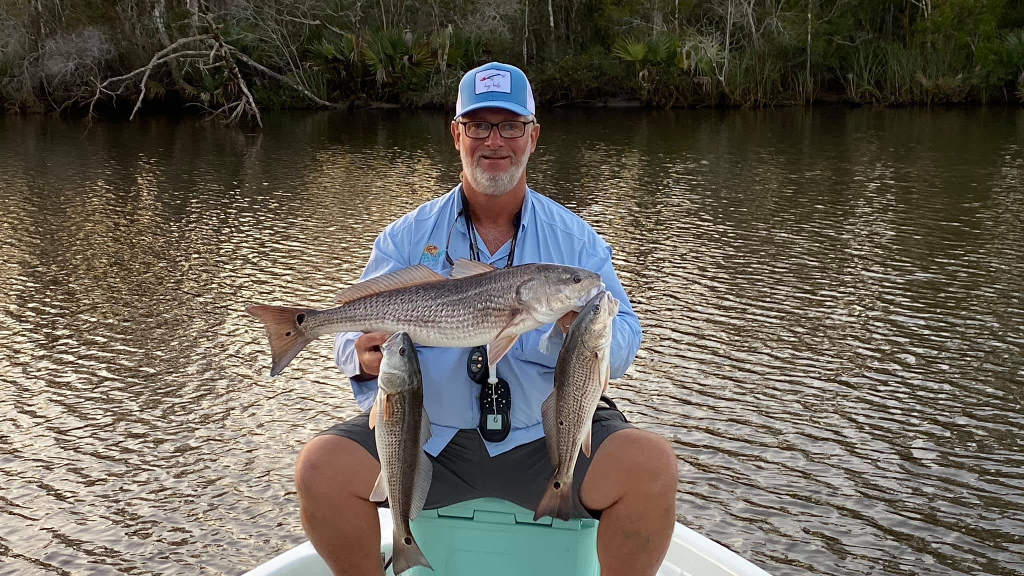 Man on boat holding three fish by a riverbank.