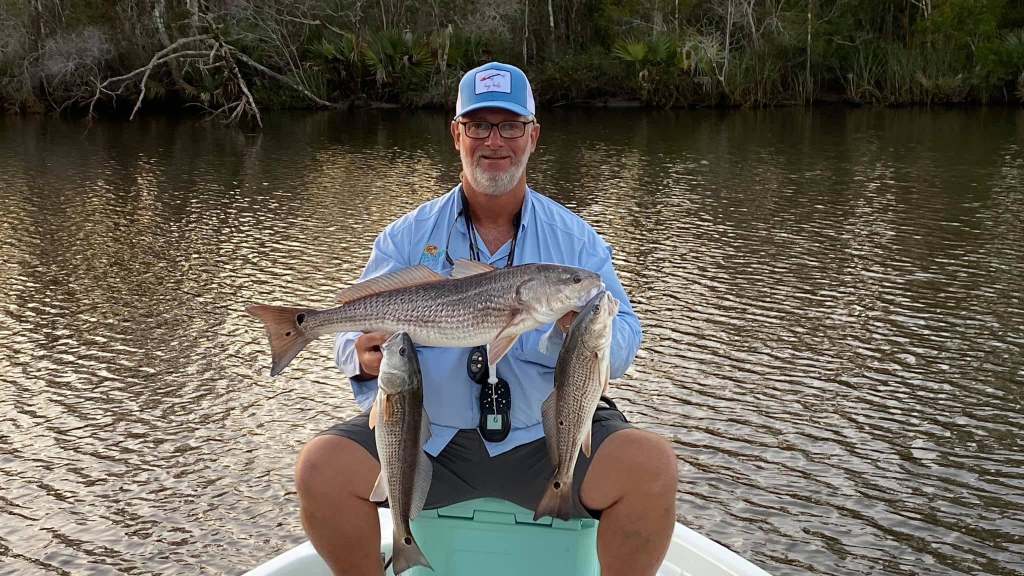 Man on boat holding three fish by a riverbank.