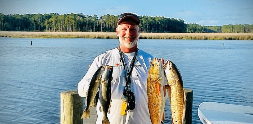 Man on dock holding two fish with a lake and forested shore in background.