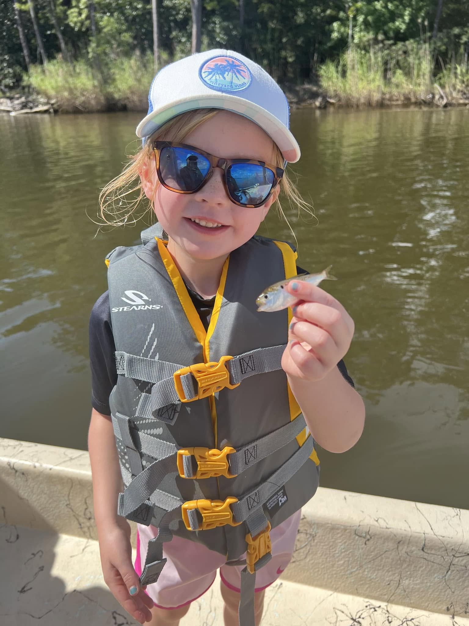 Child in a life vest holding a small fish on a boat by a river.