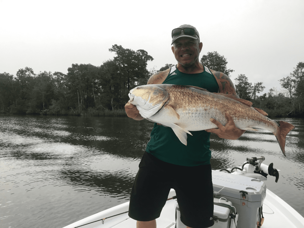 Person on boat holding large fish in front of a river with trees.